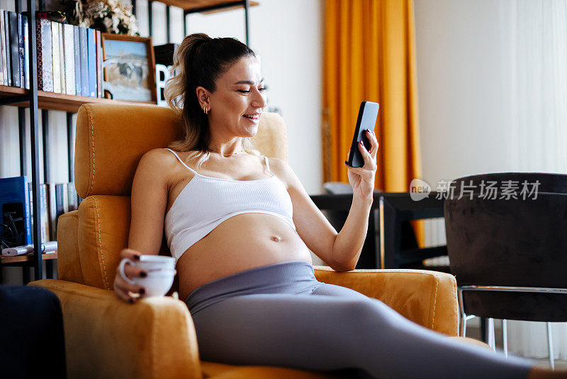 美丽的孕妇喝咖啡/茶，在家里玩手机。